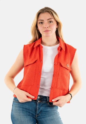 Gilet sans manches orange en tissu texturé, avec deux poches avant et une fermeture à bouton. Porté par-dessus un top côtelé blanc et un jean bleu.