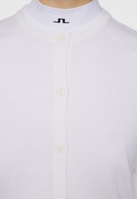 Weißer gestrickter Cardigan mit Rundhalsausschnitt, ausgestattet mit vier Knöpfen und einem kleinen, aufgenähten Logo auf der Brust. Weicher Stoff, slim fit.