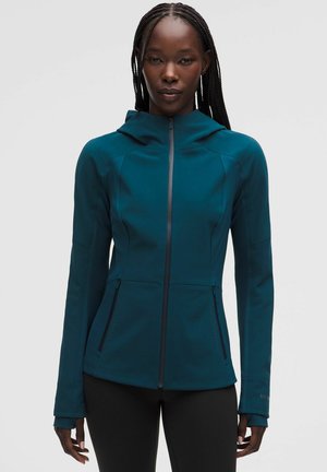 Mujer con el pelo trenzado que lleva una sudadera con capucha y cremallera en color verde azulado con bolsillos delanteros y pantalones negros, posando frente a un fondo liso de color gris claro.