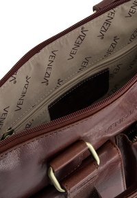 Brun lædertaske med guldspænde, åben lynlås der viser beige indvendig foring trykt med "VENEZIA" tekstmønster.