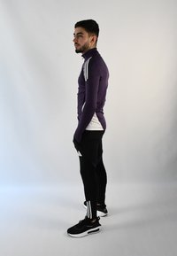 Una camiseta atlética de manga larga morada presenta un cuello alto y paneles laterales blancos; combinada con leggings negros y zapatillas negras con acentos blancos.