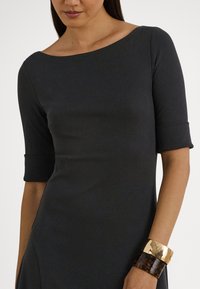 Melna pieguļoša kleita ar plašu izgriezumu un trīs ceturtdaļu piedurknēm. Akcentēta ar zelta un bruņurupuča čaulas aproci. Gluda tekstūra.