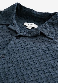 Chemise boutonnée texturée bleu foncé avec col pointu et une étiquette de la marque "NEXT" à l'intérieur de l'encolure.
