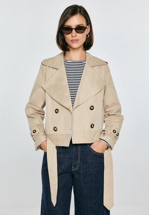 Kobieta w beżowym, dwurzędowym, krótkim trenczu, w paskowaną koszulę, ciemne dżinsy, okulary przeciwsłoneczne, z rękami w kieszeniach.