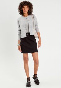 Gilet en tricot gris sur un haut gris, associé à une jupe noire. Chaussures noires avec détail en chaîne et chaussettes transparentes à pois. Design simple.