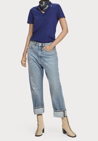 Blå v-hals t-shirt, slidte lyseblå jeans med rullede manchetter, beige ankelstøvler med sorte og mønstrede detaljer, og sort tørklæde.