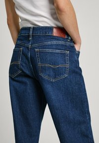 Džíny z denimu v tmavě modré barvě, s vysokým pasem, zadními kapsami s křivkovým prošitím a hnědou koženou etiketou na pase.