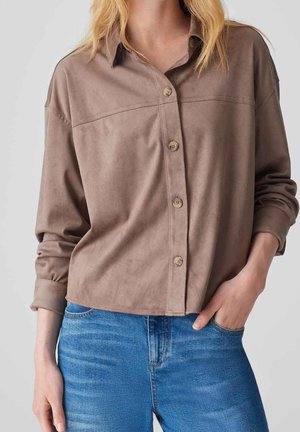 Femme portant une chemise marron ample boutonnée avec les manches retroussées et un jean bleu, une main dans la poche, sur un fond clair uni.