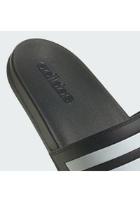 Black slide sandal with a textured upper, featuring a pronounced logo and silver stripes, crafted from synthetic materials.