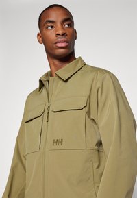 Veste zippée vert olive avec un col, deux poches poitrine et un logo brodé à l'avant. Tissu lisse avec une finition mate.