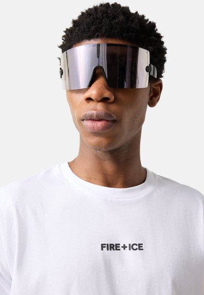 Jeune homme portant de grandes lunettes de soleil à visière réfléchissante et un T-shirt blanc avec le texte "FIRE+ICE" sur la poitrine, sur un fond clair.