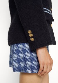 Manche de blazer texturée noire avec trois boutons dorés en forme d'ancre, associée à une jupe ou un short à motif pied-de-poule bleu et blanc.