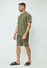 Chemise à manches courtes côtelée vert olive et shorts assortis, avec un col en V et une fermeture à boutons. Portée avec des sandales noires à enfiler.
