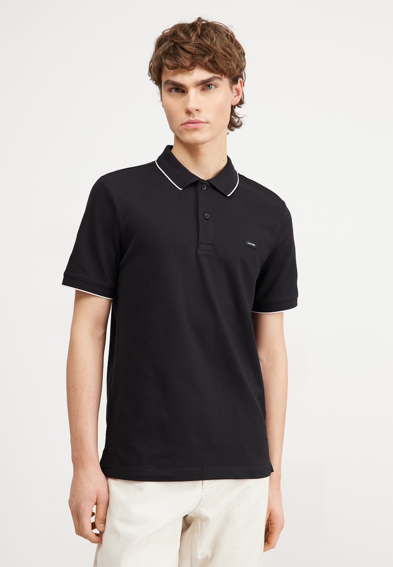 Black polo shirt made of cotton with a classic collar, two button placket, and white piping on the collar and sleeves. Small logo on chest.