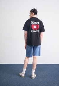 Zwart t-shirt met witte tekst en een rood hartgrafiek op de achterkant, gecombineerd met denim shorts en grijze sandalen. Soepele stof, casual pasvorm.