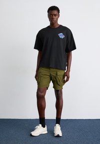 Black oversized t-shirt with colorful graphic, olive green cargo shorts, and white sneakers. Man stands against a plain background.
