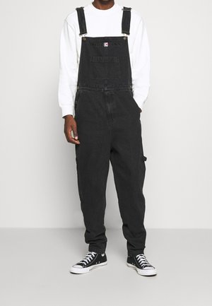 Personne portant une salopette en denim noire avec poche avant et logo "KARL KANI", chemise blanche à manches longues et baskets montantes noires, debout.
