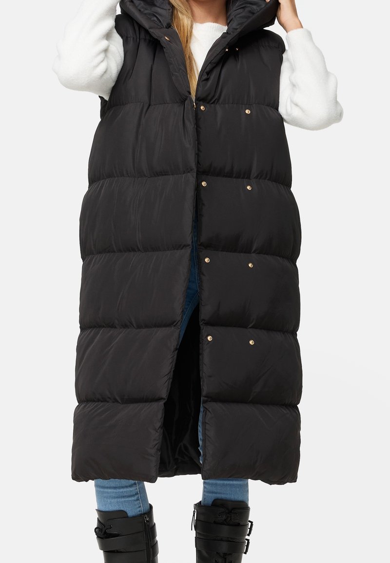 Long gilet matelassé noir avec capuche, doté de boutons-pression dorés et d'une texture lisse et brillante. Porté par-dessus un pull blanc et un jean bleu.