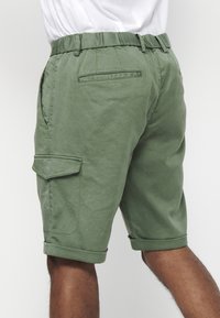 Grüne Baumwoll-Cargo-Shorts mit umgeschlagenen Säumen, seitlichen Taschen und einer Gesäßtasche mit quadratischem Klappdetail. Glatte Textur mit einem lässigen Schnitt.