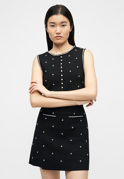 Γυναίκα με μακριά σκουρόχρωμα μαλλιά που φοράει αμάνικο μαύρο φόρεμα διακοσμημένο με κουμπιά που μοιάζουν με μαργαριτάρια και μικρές λουλουδάτες διακοσμήσεις, με σταυρωμένα χέρια.