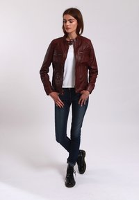 Burgundy leather jacket with a fitted design, round collar, zippered pockets, and textured finish, worn over a white top and blue jeans.
