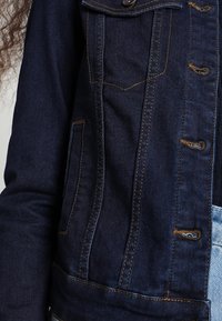 Tmavě modrá džínová bunda s knoflíkovou přední částí, dvěma náprsními kapsami a kontrastními oranžovými švy. Má klasický límec a strukturovaný střih.