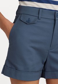 Modré látkové šortky s hladkou texturou, předními kapsami a detailem složeného lemu. Zapínají se na dvě modré knoflíky na každé kapse.