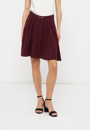 Femme portant une jupe plissée bordeaux longueur genou avec un détail boucle en métal, un haut blanc et des sandales à talons noires avec bride à la cheville.