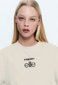 Camiseta beige con escote redondo que presenta texto impreso en negro "FREDDY elite" en una fuente moderna. Textura suave, corte relajado.
