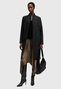 Cappotto lungo nero, top nero texturizzato, gonna plissettata dorata e stivali neri a punta fino al ginocchio, con una borsa nera texturizzata.
