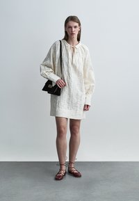 Λευκό φόρεμα με υφή, με σούρα στο ντεκολτέ και λεπτομέρεια δεσίματος, συνδυασμένο με μαύρη τσάντα χιαστί και κόκκινες δαντελωτές μπαλαρίνες με κορδόνια.