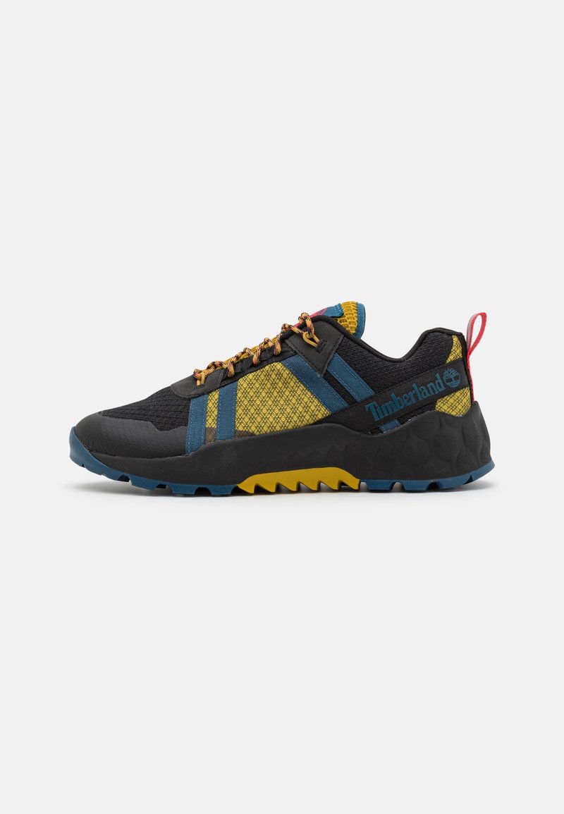 Baskets basses de sport avec dessus noir, jaune et bleu, tissu à motifs, semelle robuste bleu et jaune, et logo Timberland sur le côté.
