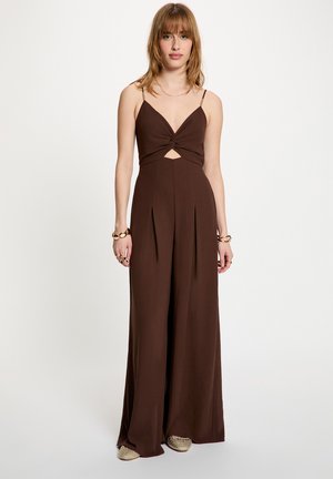 Nuori nainen, jolla on päällään ruskea hihaton jumpsuit, jossa edessä kierre ja leikkaus, asusteina rannekoruja ja sormuksia, seisoo valkoisella taustalla.
