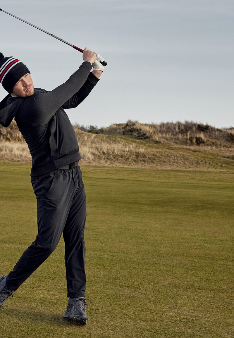 Golfer in einem schwarzen Hoodie, schwarzen Hosen und grauen Schuhen, der einen Schläger auf grünem Gras schwingt, mit einer Landschaft aus Hügeln und einem bewölkten Himmel.