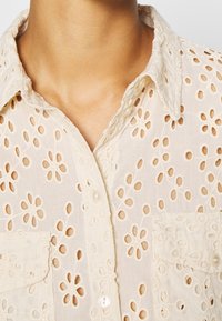 Camicia beige chiaro con motivo floreale a occhielli, chiusura con bottoni sul davanti e due tasche frontali. Tessuto morbido e strutturato con un delicato colletto.