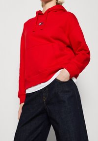 Czerwona bluza z kapturem, z kieszenią typu kangur, ściągaczami przy rękawach i małym logo na piersi. Nosi się ją na białej koszulce, zestawiona z ciemnymi dżinsami.