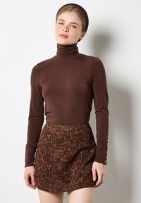 Młoda kobieta w brązowym swetrze z golfem, mini spódnicy w wzór lamparta i pasującej brązowej opasce na głowę, stojąca przy białej ścianie.