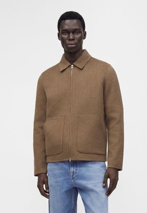 Homme portant une veste marron zippée avec col et grandes poches avant, associée à un jean bleu clair, debout devant un fond blanc uni.