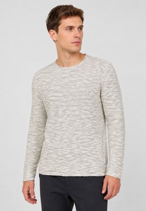 Jonge man die een lichtgrijze gestructureerde lange mouw trui en een donkere broek draagt, kijkt iets naar rechts tegen een effen achtergrond.