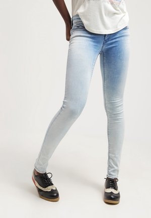 En person iført lyseblå stramme jeans og sort-hvide snørebåndssko, stående med det ene ben bøjet fremad.