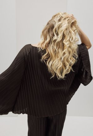 Pikkade lokkis blondide juustega naine, kes kannab lahtist musta plisseeritud ülaosa ja pükse, käsi juustel, näoga selja poole lihtsa tausta ees.