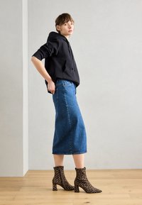 Sweatshirt à capuche noir, jupe midi en denim bleu, et bottines à talons blocs en imprimé léopard marron. Sol en bois et arrière-plan neutre.