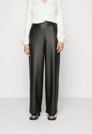 Vrouw in hoge taille zwarte nepleer broek met wijde pijpen, witte korte cardigan en metallic platform schoenen op een effen achtergrond.