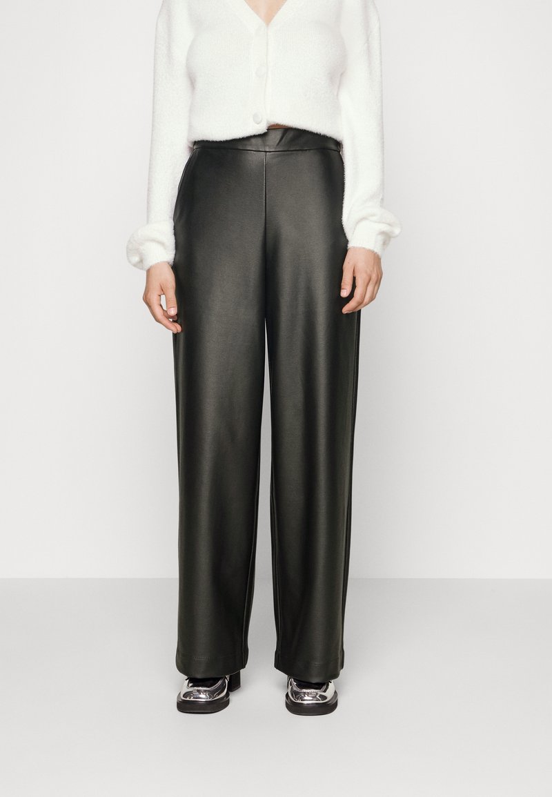 Kvinna som bär högmidjade svarta fusk läder vida byxor, vit kortärmad kardigan och metalliska plattformsskor mot en neutral bakgrund.