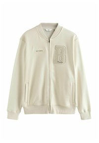 Lys beige jakke med lynlås, ribbet krave, sidelommer og broderet tekst "Williamsburg NY STATE", samt en tekstureret "B" patch.