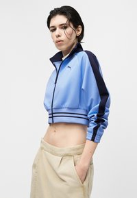 Νεαρός με κοντά σκούρα μαλλιά που φοράει ανοιχτό μπλε κοντό μπουφάν με σκούρες ρίγες και μπεζ παντελόνι, στέκεται με το ένα χέρι στην τσέπη.