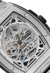 Zilveren roestvrijstalen horloge met een skeleton ontwerp, dat mechanische componenten, tandwielen en paarse accenten toont. Zwarte gestructureerde band.