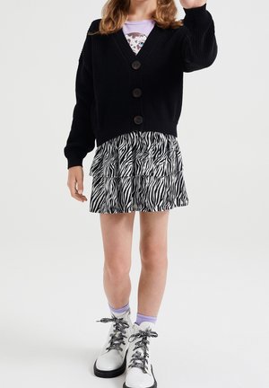 Młoda osoba ubrana w czarny rozpinany sweter, spódnicę w zebrę, białe buty z sznurowadłami w zebrę oraz jasnofioletowe skarpetki, stojąca na tle białego tła.