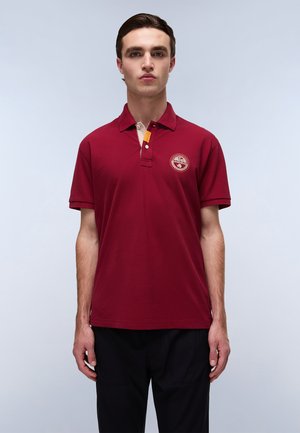 Burgundy polo shirt med krave med to knapper, korte ærmer og en gul detalje ved halsen. Har et cirkulært broderet logo på brystet.