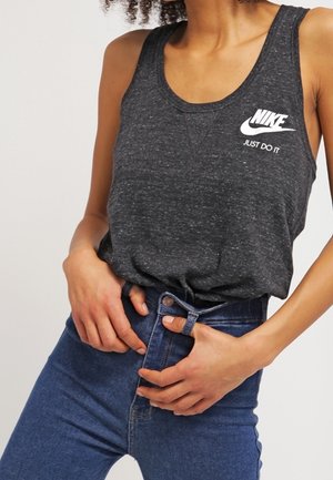 Vrouw draagt een donkergrijze Nike tanktop met wit logo en slogan, ze doet een hooggetailleerde blauwe spijkerbroek dicht.